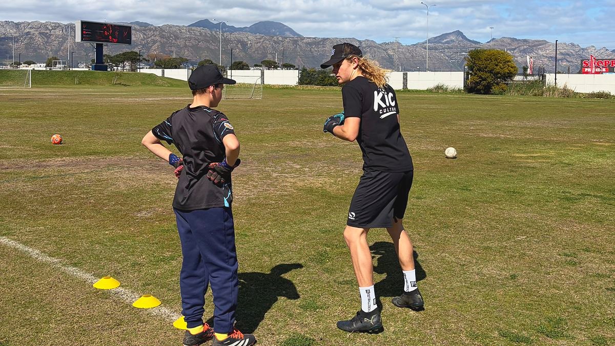 Jack running a goalkeeping drill with a young keeper in gloves, mountains behind the field.